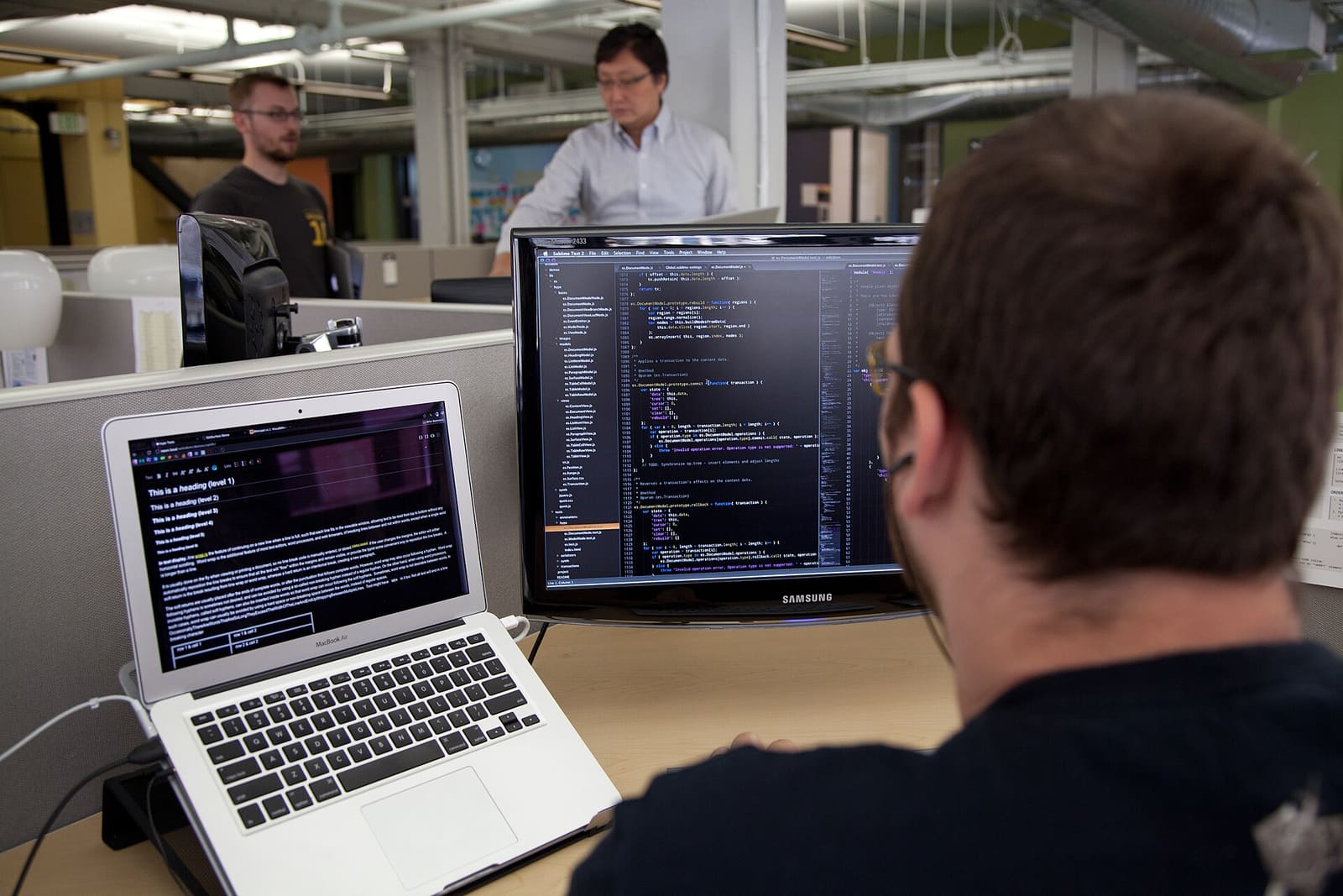 A developer at a desk looking at a screen full of code, with a laptop open beside him