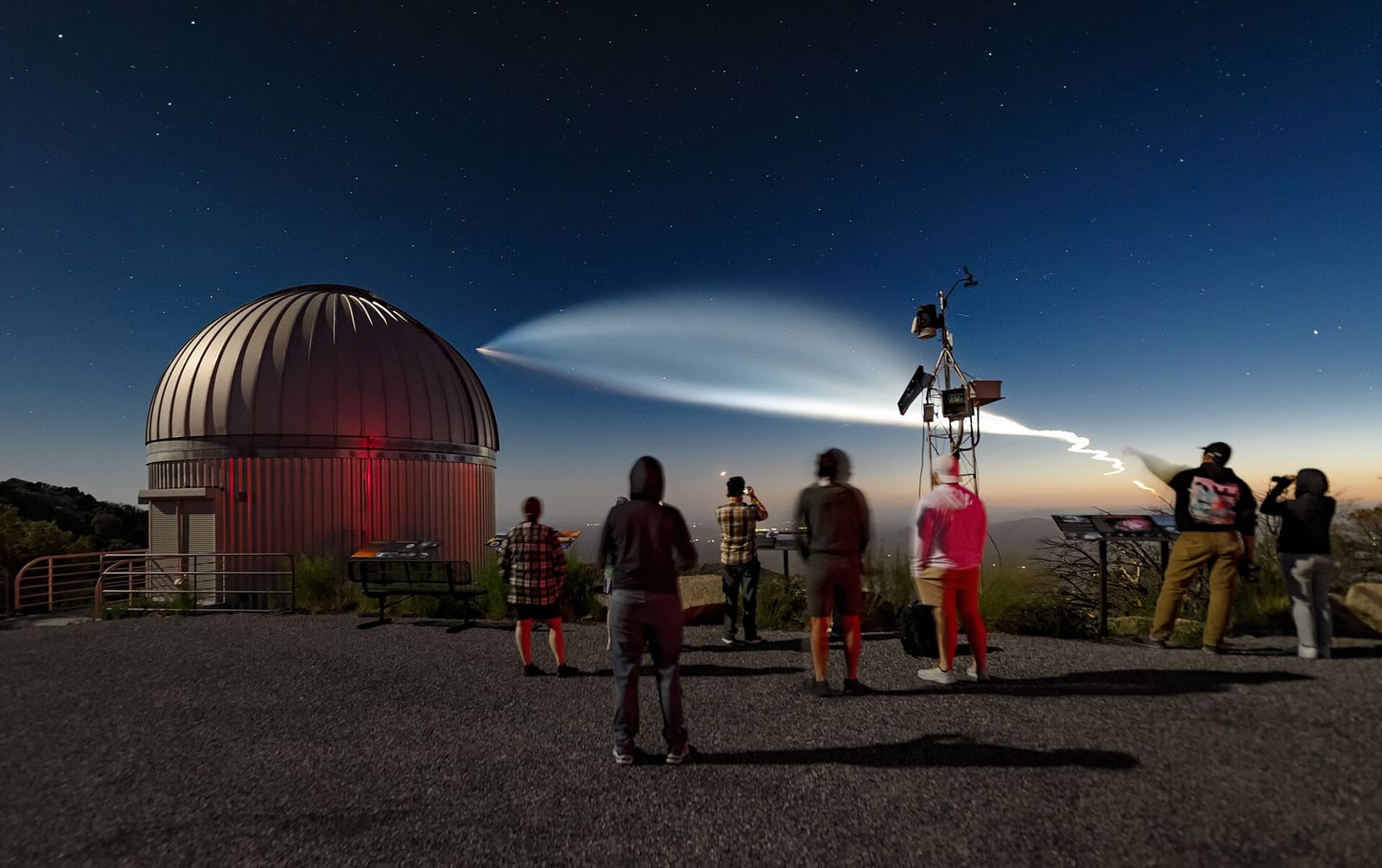 A SpaceX Falcon 9 launch trail glowing in the twilight sky above Kitt Peak National Observatory, with visitors watching