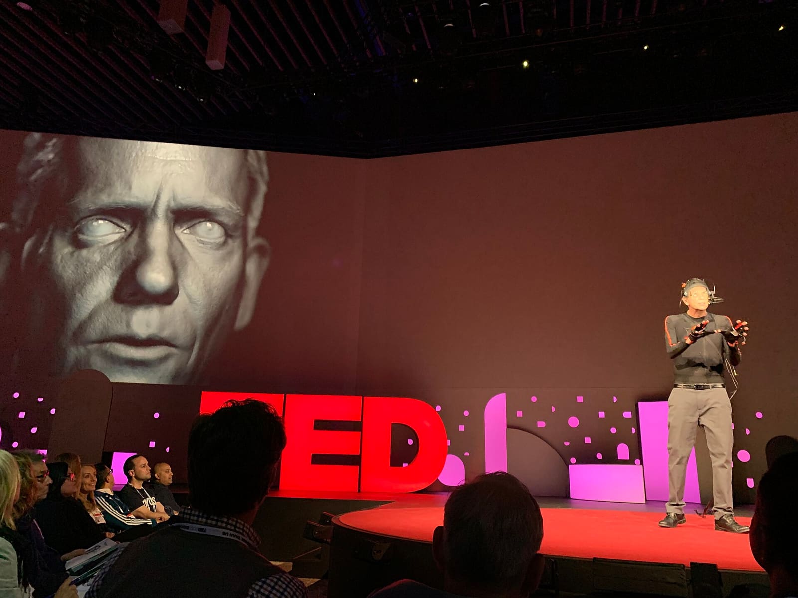 A stage screen showing a computer-generated digital human face next to the real person